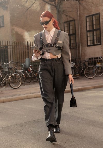Blazer gris corto con correas de arnés, camisa blanca, jeans oscuros y botas negras hasta el tobillo. Los accesorios incluyen gafas de sol y un bolso pequeño.