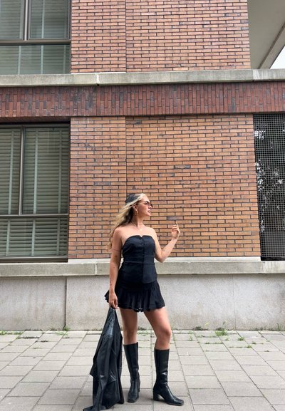 Vestido negro sin tirantes con un dobladillo con volantes, corsé con botones. Lleva botas negras hasta la rodilla y sostiene una chaqueta de cuero negra.