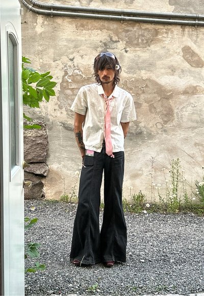 Camisa blanca de manga corta con una corbata rosa, pantalones vaqueros de pierna ancha negros y gafas de sol; de pie sobre grava con plantas al fondo.