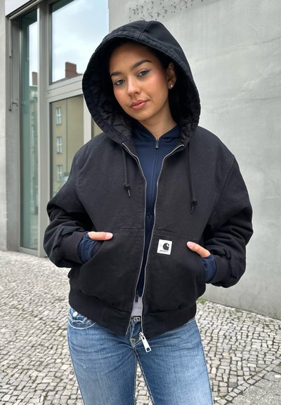 Persona joven con chaqueta negra con capucha y manos en los bolsillos, de pie junto a una pared de hormigón y una ventana en una acera adoquinada.