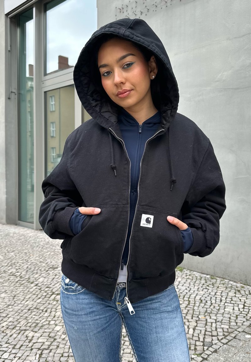 Jeune personne portant une veste noire à capuche, les mains dans les poches, debout près d'un mur en béton et d'une fenêtre sur un trottoir pavé.