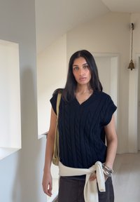 Haut noir à manches courtes en maille torsadée avec un col en V, associé à un pull crème noué à la taille, et un pantalon marron ; sac vert et bracelets argentés.