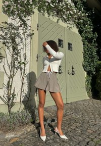 Cardigan blanc à côtes, mini-jupe plissée beige avec cordon de serrage et chaussures �à talons blanches, devant une porte verte et de la végétation.