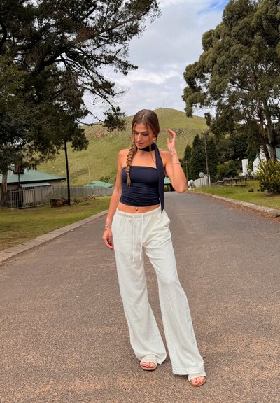 Joven con el cabello trenzado, vestida con top negro sin tirantes y pantalones blancos de pierna ancha, de pie en una carretera tranquila con vegetación y colinas al fondo.