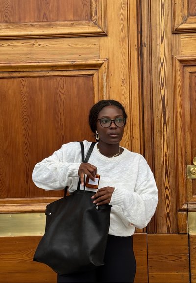 Mujer con gafas y suéter blanco sosteniendo una bolsa tote negra, colocando una pequeña botella marrón en su interior, de pie frente a puertas de madera.