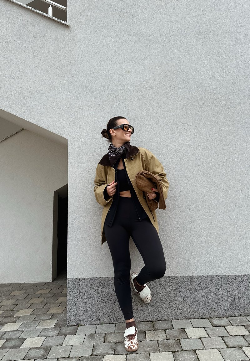 Femme en leggings noirs et crop top, veste beige et écharpe, tenant un sac marron, debout sur un pavé devant un mur gris, souriant et regardant de côté.