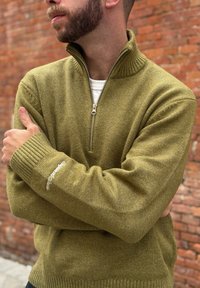 Homme à la barbe taillée portant un pull zippé vert olive avec une manche brodée, debout les bras croisés devant un mur en briques.