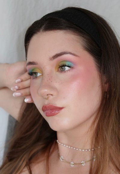 Primer plano de una persona con cabello castaño claro y una diadema negra, que exhibe un maquillaje de ojos colorido, un piercing en el septum de oro y collares de perlas.