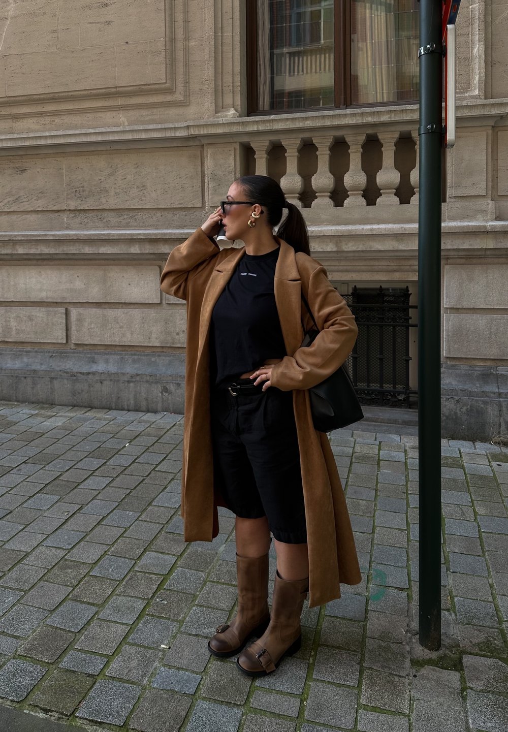 Cappotto lungo marrone sopra una maglietta nera e pantaloni corti neri; stivali marroni alti fino al ginocchio con fibbie; borsa nera; in piedi su una strada ciottolosa.