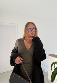 Top verde a coste con scollo a V profondo e cappotto nero oversize. Borsa nera con dettagli marroni. Occhiali blu vivace. Ambientazione interna semplice.