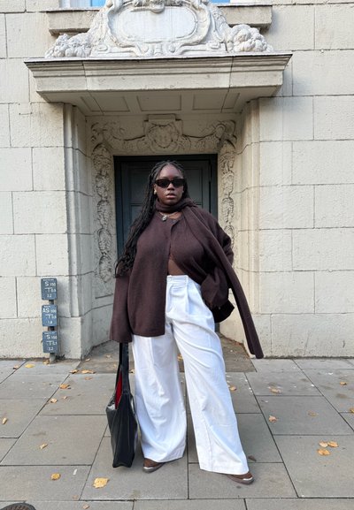Cárdigan de lana marrón de gran tamaño sobre una blusa corta, pantalones anchos blancos, gafas de sol negras y un bolso negro, todo frente a una pared texturizada.