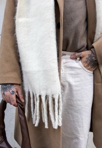 Hnedý vlnený kabát, béžový rebrovaný sveter, krémový šál s strapcami, svetlá sukňa a hnedá kabelka, s viditeľnými zložitými tetovaniami na ruke.