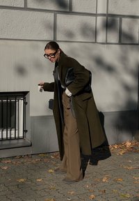 Μακρύ ελαιοπράσινο παλτό πάνω από ένα γκρίζο τοπ, φαρδιά μπεζ παντελόν�ια και γκρίζα παπούτσια. Μαύρη τσάντα και κρατάει ένα μικρό αντικείμενο. Ηλιασμένο αστικό σκηνικό.