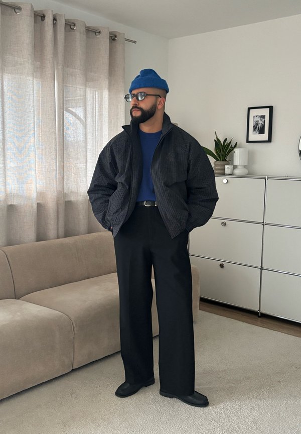 Un homme barbu portant un bonnet bleu, des lunettes, une veste sombre et un pantalon à jambes larges se tient à côté d’un canapé dans un salon.