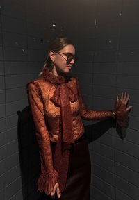 Csipkefelső égett narancssárga színben, fodros gallérral és hosszú ujjakkal, hozzáillő megkötővel és áttetsző virágmintával; sötét szoknyával párosítva.