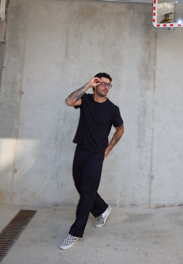 Hombre con tatuajes vestido con ropa negra y zapatos a cuadros ajusta sus gafas mientras está apoyado contra una pared de cemento cerca de un espejo de seguridad.