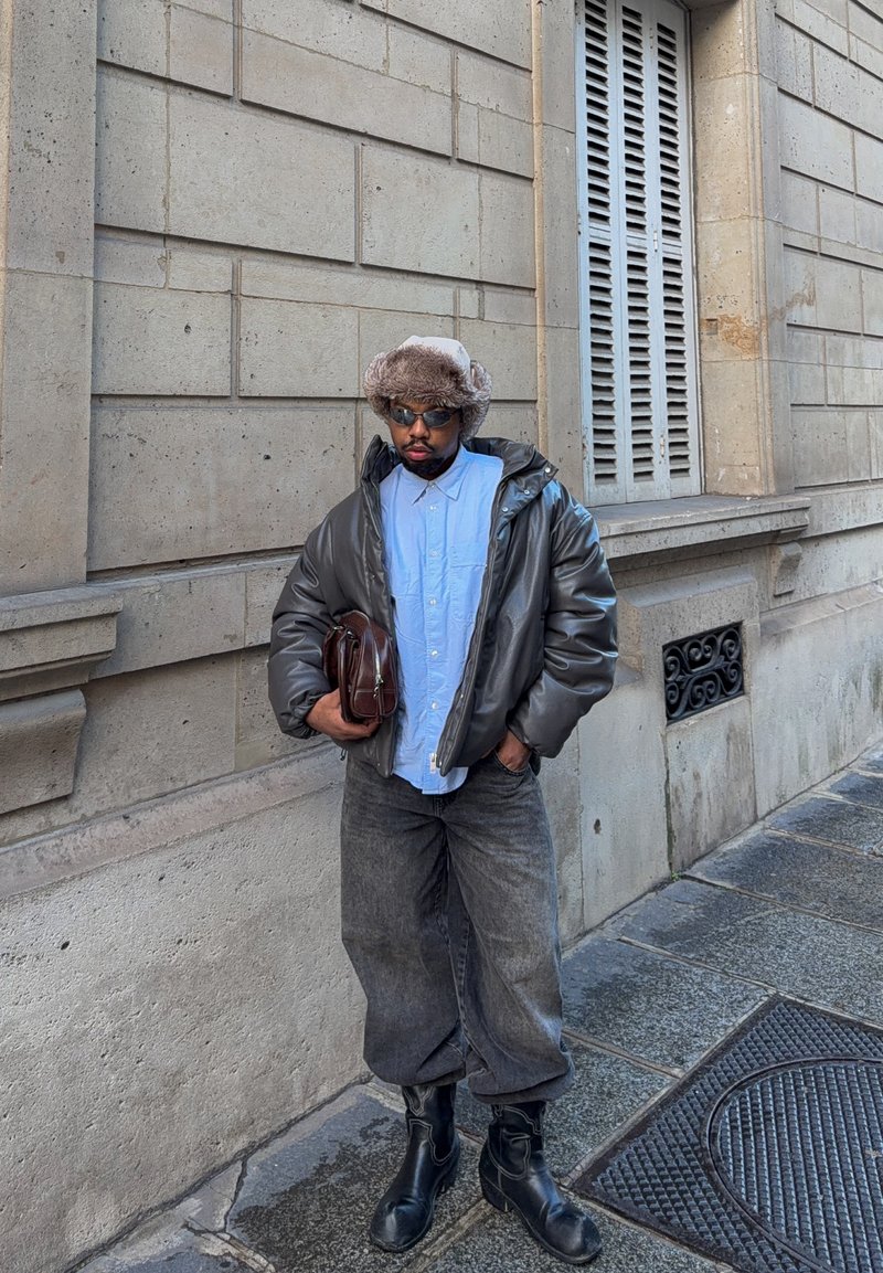Graue Steppjacke, hellblaues Hemd, übergroße dunkle Jeans, schwarze Leder-Stiefeletten, braune Tasche und Pelzmütze; vor einer Steinmauer stehend.