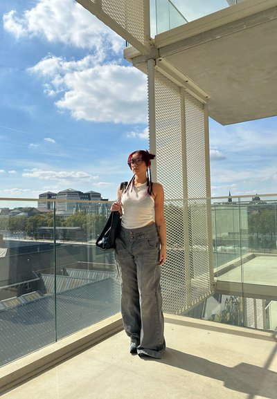 Modelo vistiendo una blusa sin mangas de color gris claro y jeans amplios grises con bolsillos frontales. Sosteniendo un bolso tote negro, de pie en una terraza.