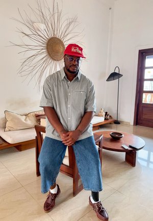 Hombre con gafas y gorra roja sentado en una silla de madera con pierna protésica, camisa de rayas, vaqueros azules en una sala de estar luminosa con decoración y muebles.