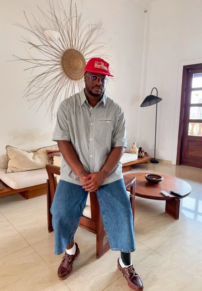 Hombre con gafas y gorra roja sentado en una silla de madera con pierna protésica, camisa de rayas, vaqueros azules en una sala de estar luminosa con decoración y muebles.