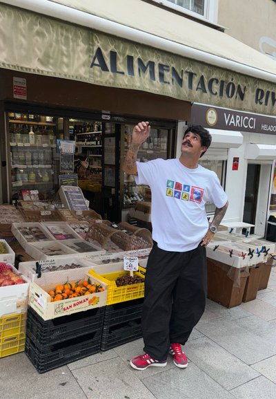Camiseta blanca con un gráfico colorido en la parte frontal, pantalones negros y zapatillas rojas. Mercado al aire libre con cajas de madera de frutas y productos visibles.