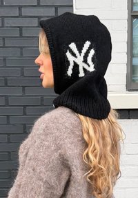 Femme aux longs cheveux blonds ondulés portant une cagoule en tricot noire avec un logo blanc, face à gauche, debout contre des murs en briques gris et blanc.