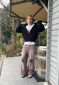 Jeune personne aux cheveux bouclés ajustant un pull zippé noir, portant un pantalon large marron déchiré, debout à l'extérieur à côté d'un tonneau en bois et d'un jardin.