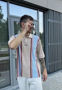 Kortærmet polo shirt med struktureret strik i beige, lyseblå og røde striber; har en krave og logo-detalje. Modellen bærer solbriller.