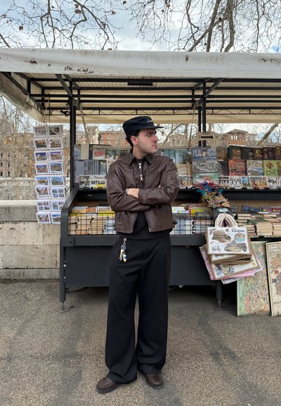 Hombre con chaqueta de cuero marrón y gorra negra, de pie con los brazos cruzados frente a un puesto de libros al aire libre que vende mapas, postales y libros.