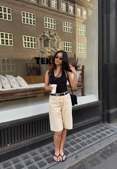 Camiseta negra sin mangas, pantalones cortos beige, cinturón negro y chanclas. Sosteniendo una taza de café, de pie frente a un escaparate de cristal.