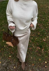 Pull en tricot crème à manches larges, jupe beige semblable à de la soie, sac à main en daim marron avec un charm en peluche, et escarpins nude pointus sur un fond herbeux.