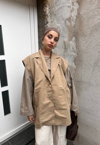 Blazer oversize beige con ampie rever e bottoni argentati, indossato sopra una maglia grigia; abbinato a pantaloni beige chiaro e una borsa a tracolla marrone.