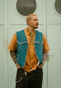 Veste en jean à bords effilochés portée sur une chemise à manches courtes rayée orange. Les accessoires comprennent une écharpe verte et un porte-clés avec des perles.