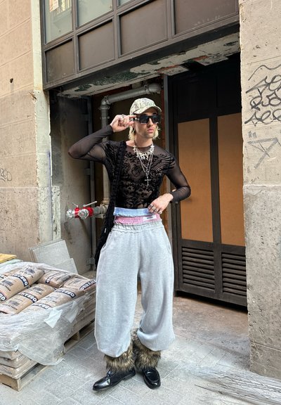 Top de malla negro de manga larga con diseño floral, pantalones de chándal grises con borde de piel, zapatos negros, cadenas en capas y una gorra con patrón.