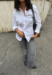 Vrouw in een witte blouse, grijze jeans en zwarte schoenen die op een stoep staat naast een gestructureerde muur, met een bruine tas op haar schouder.