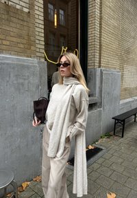 Camisola de malha cinza claro com gola alta e detalhe de lenço drapeado. Combinada com calças largas de cor creme e acessórios como óculos de sol escuros e uma pequena mala.