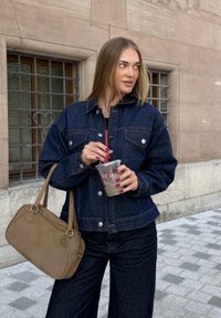 Donkerblauwe denimjack met zilveren knopen, gecombineerd met wijde jeans. Houdt een heldere beker met een rietje vast en een tan handtas.