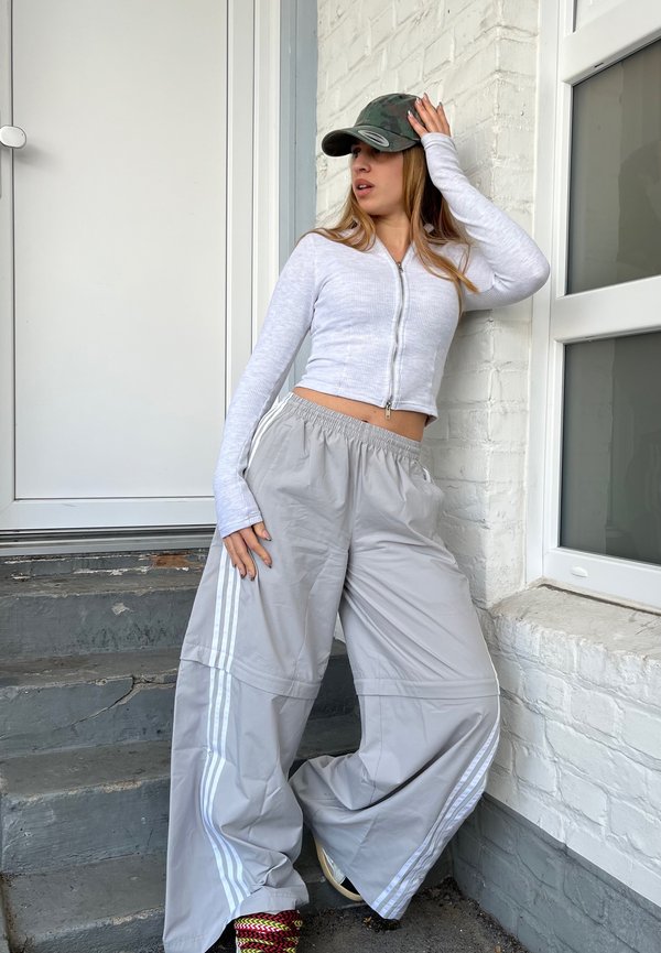 Pantalones de pierna ancha gris claro con rayas blancas. Parte superior ajustada blanca con cremallera. Gorra de camuflaje. La modelo posa frente a una pared de ladrillos blancos, con una escalera visible.