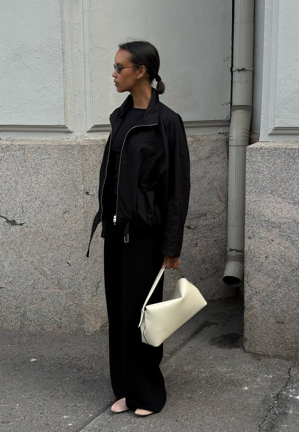Mujer con chaqueta y pantalones negros, sosteniendo un bolso blanco, con gafas de sol, de pie frente a una pared blanca y de piedra gris.