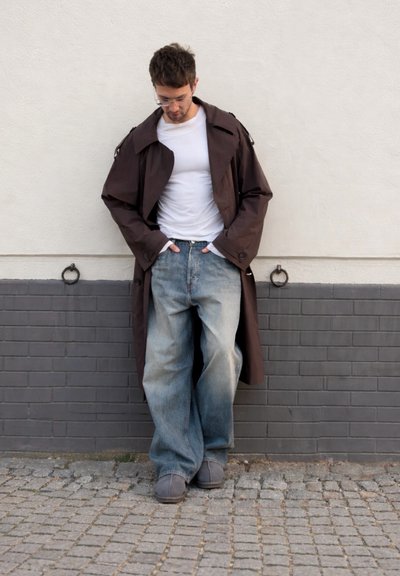 Joven con gafas que lleva un abrigo largo oscuro, camisa blanca, vaqueros azules holgados y zapatos grises, de pie junto a una pared de ladrillos blanca y gris.