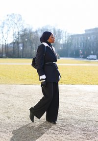 Femme en hijab noir, blazer ajusté, pantalon large et gants, marchant en plein air avec un sac à dos dans un parc par une journée ensoleillée.