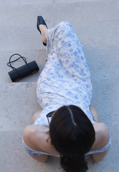 Vestido blanco con patrón floral azul; bolsa cilíndrica negra con dos tirantes delgados al lado. Zapatos de punta negra sobre una superficie texturizada.