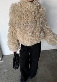 Personne portant une veste en fausse fourrure bouclée beige et un pantalon noir ample, tenant un sac à main en cuir noir devant un mur uni.
