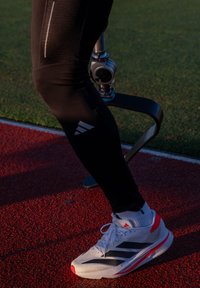 Valge Adidas jalanõu punaste detailidega, kolm musta triipu, mustade retuusidega. Taustal on nähtav tera protees.