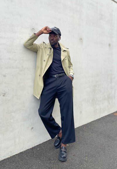 Gabardina beige con grandes botones, combinada con una camiseta negra y pantalones. Zapatos negros con suelas gruesas y detalle de cadena.