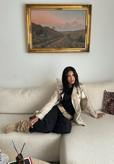 Gabardina beige, blusa negra y pantalones negros holgados. Sofá crema con textura y cojín con estampado de leopardo. Bolso marrón con detalles decorativos.