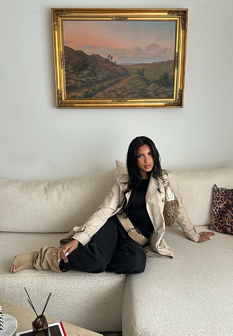 Trench beige, top nero e pantaloni neri morbidi. Divano crema con cuscino a stampa leopardata. Borsa marrone con dettagli accentuati.