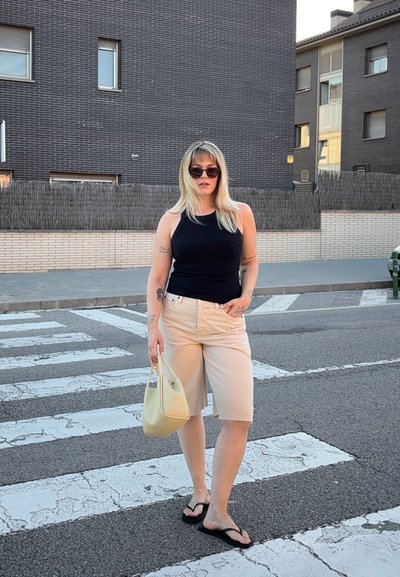 Mujer de pelo rubio, gafas de sol, camiseta de tirantes negra, pantalones cortos beige y chanclas sostiene un bolso color crema mientras está de pie en un paso de peatones en una ciudad.