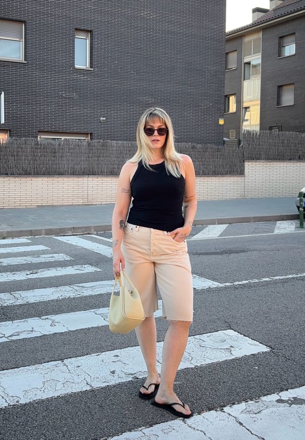 Vrouw met blond haar, zonnebril, zwart hemdje, beige korte broek en slippers houdt een crèmekleurige handtas vast terwijl ze op een zebrapad in een stad staat.