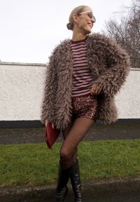 Manteau en fausse fourrure marron, chemise rayée bordeaux et blanche, short pailleté, collants transparents à motifs, et bottes noires montant jusqu'aux genoux. Tient une pochette rouge.